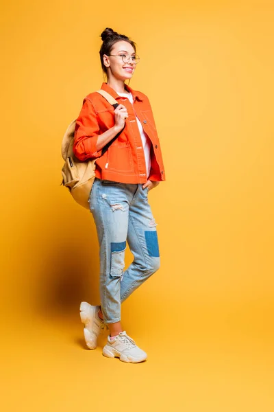 Вид улыбающегося студента в полный рост с рюкзаком, отводящим взгляд, держа руку в кармане на желтом фоне — стоковое фото