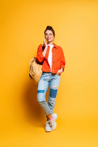 Full length view of cheerful student with backpack talking on smartphone while holding hand in pocket on yellow background — Stock Photo