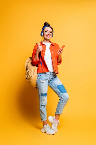 Estudante sorrindo atraente em fones de ouvido segurando mochila e smartphone enquanto olha para a câmera no fundo amarelo — Fotografia de Stock