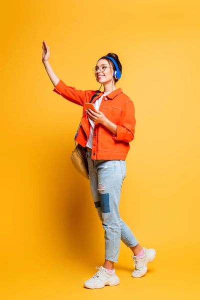 Vista completa de estudiante alegre en auriculares agitando la mano mientras mira hacia otro lado en el fondo amarillo - foto de stock