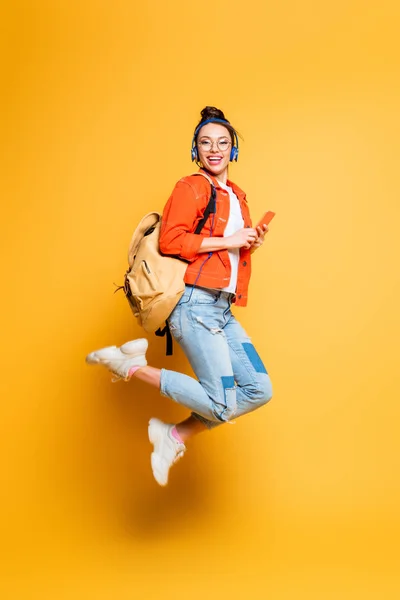 Excited student in headset and glasses jumping while holding smartphone on yellow background — Stock Photo