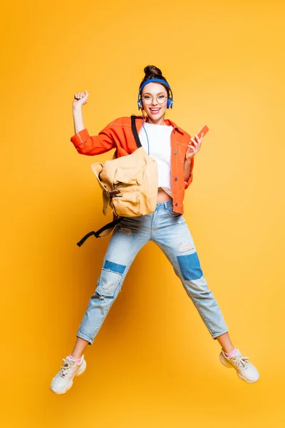 Cheerful student in headset and glasses jumping while holding smartphone on yellow background — Stock Photo