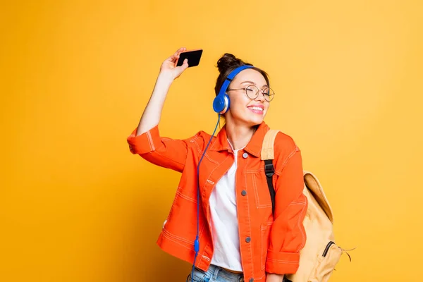 Estudiante alegre en auriculares y gafas que sostienen el teléfono inteligente sobre fondo amarillo - foto de stock
