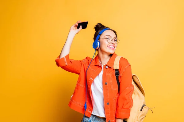 Studente allegro in auricolare e occhiali che tengono smartphone su sfondo giallo — Foto stock