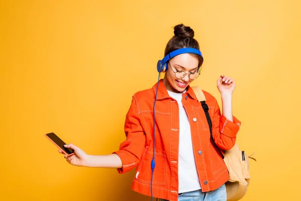 Cheerful student in headphones holding smartphone on yellow background — Stock Photo