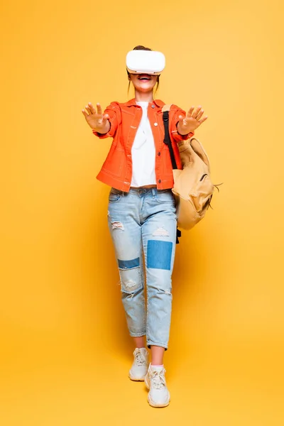 Ragazza eccitata con zaino in piedi con le mani tese durante l'utilizzo di auricolare vr su sfondo giallo — Foto stock