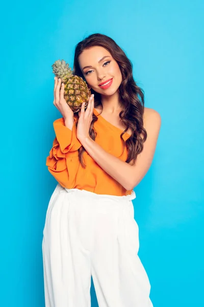 Atractiva y sonriente chica mirando a la cámara mientras sostiene la piña madura aislada en azul - foto de stock