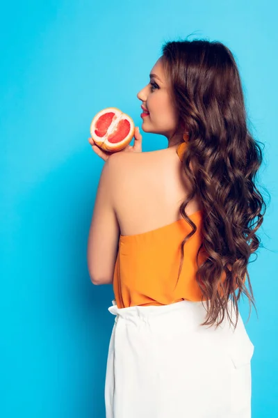 Attractive long-haired girl holding half of fresh grapefruit on blue background — Stock Photo