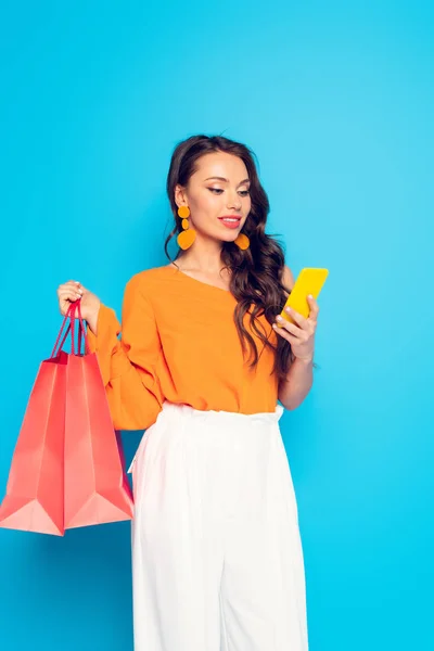 Attraktive trendige Mädchen plaudert auf dem Smartphone, während sie Einkaufstüten auf blauem Hintergrund hält — Stock Photo