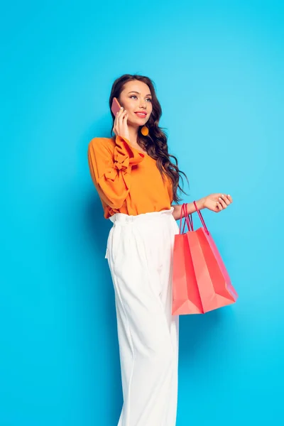 Atractiva chica sonriente hablando en el teléfono inteligente mientras sostiene bolsas de compras sobre fondo azul - foto de stock