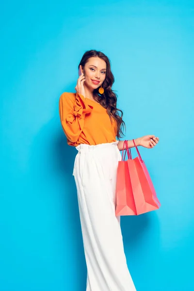 Attractive fashionable girl talking on smartphone while holding shopping bags on blue background — Stock Photo