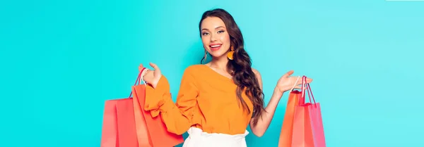 Plano panorámico de la chica de moda sonriente sosteniendo bolsas de compras y mirando a la cámara en el fondo azul - foto de stock