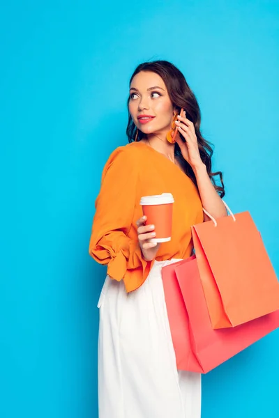 Atractiva chica elegante sosteniendo café para ir y bolsas de compras mientras habla en el teléfono inteligente de distancia en fondo azul - foto de stock