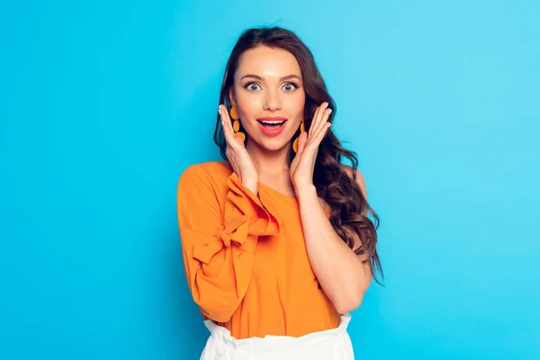 Eccitata ragazza elegante che si tiene per mano vicino al viso e guardando la fotocamera su sfondo blu — Foto stock