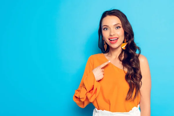 Happy stylish girl smiling at camera while pointing with finger on blue background — Stock Photo