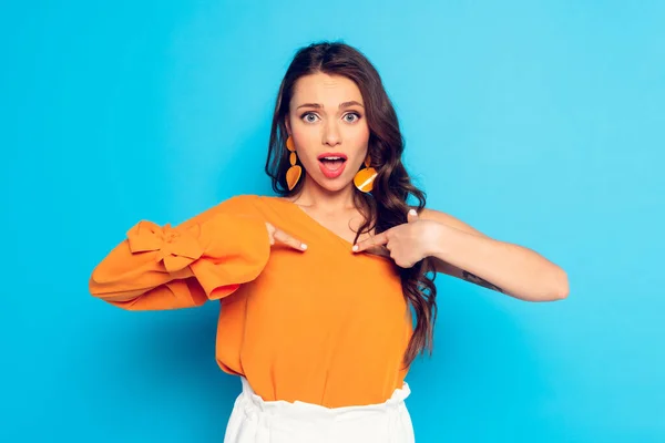 Elegant, shocked girl pointing with fingers at herself while looking at camera on blue background — Stock Photo