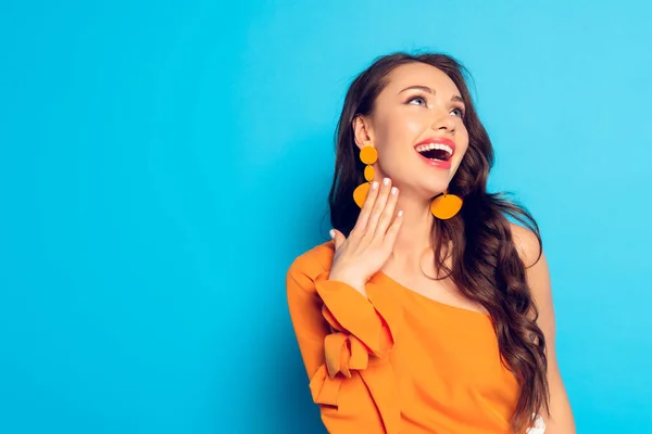 Chica de moda, emocionada mirando hacia otro lado mientras sostiene la mano cerca del cuello sobre fondo azul - foto de stock