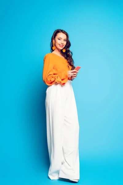 Full length view of beautiful, happy woman in stylish clothes using smartphone and smiling at camera on blue background — Stock Photo