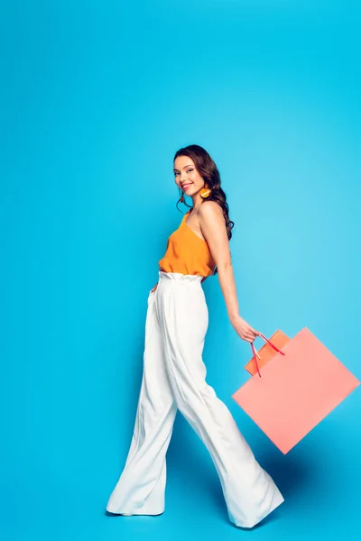 Vista completa de la mujer alegre y elegante caminando con bolsas de compras de color rosa y sonriendo a la cámara sobre fondo azul - foto de stock