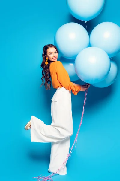 Повний вигляд щасливої стильної жінки, що стоїть на одній нозі, тримаючи великі святкові кульки на синьому фоні — стокове фото