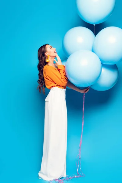 Erstaunt stilvolle Frau berührt Kinn, während man auf großen festlichen Luftballons auf blauem Hintergrund — Stockfoto