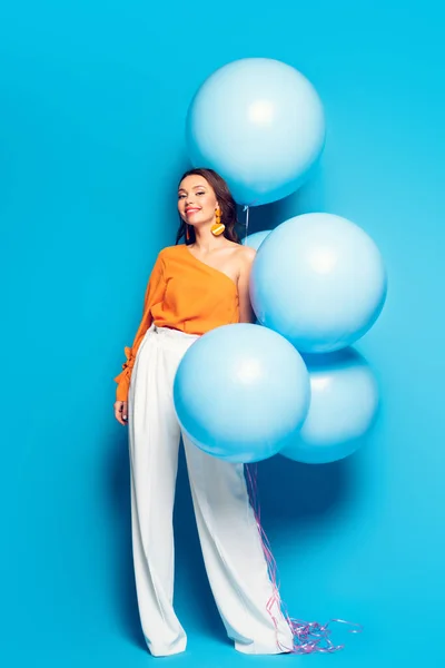 Полный вид веселой стильной женщины с большими праздничными воздушными шарами, улыбающейся в камеру на синем фоне — стоковое фото