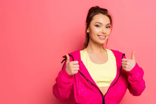 Junge fröhliche Sportlerin zeigt Daumen nach oben, während sie auf rosa Hintergrund in die Kamera lächelt — Stockfoto