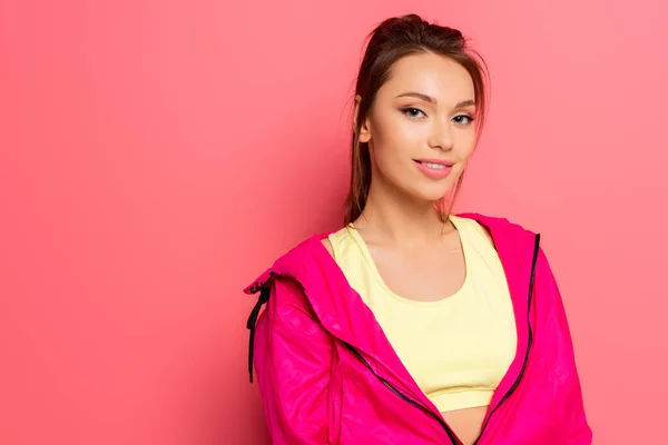 Attractive, confident sportswoman in pink windbreaker smiling at camera on pink background — Stock Photo