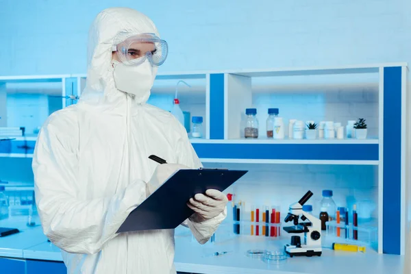 Científico en traje de materiales peligrosos y gafas sujetando el portapapeles y la pluma cerca del microscopio - foto de stock