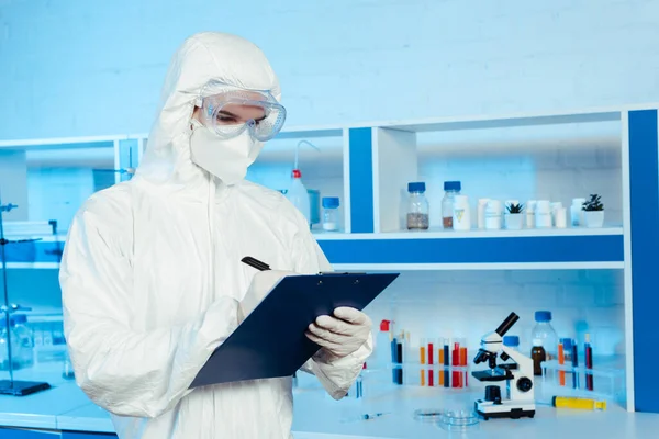 Cientista em terno hazmat e óculos escrevendo enquanto segurando prancheta perto do microscópio — Fotografia de Stock