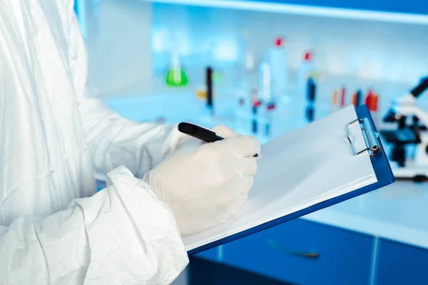 Vista recortada del científico en traje de materiales peligrosos escribiendo en papel en blanco mientras sostiene el portapapeles - foto de stock