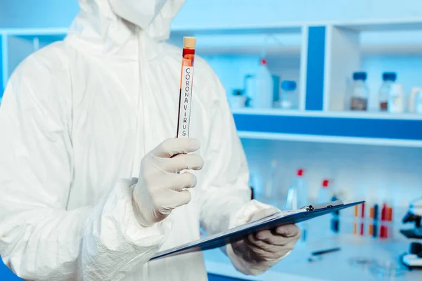 Cropped view of scientist in hazmat suit holding test tube with coronavirus lettering — Stock Photo