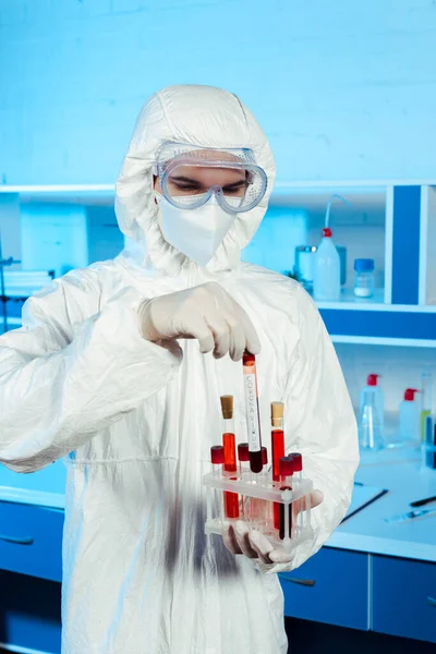 Científico en traje de materiales peligrosos y guantes de látex que sostiene el tubo de ensayo con letras de coronavirus - foto de stock