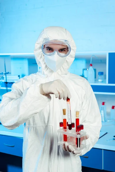 Científico en traje de materiales peligrosos y guantes de látex que sostiene el tubo de ensayo con letras - foto de stock
