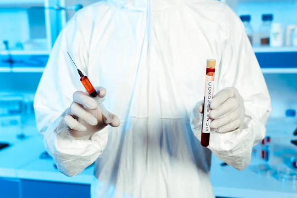 Vista recortada del científico en guantes de látex que sostienen la jeringa y el tubo de ensayo con letras de coronavirus - foto de stock