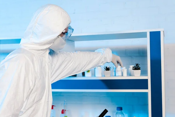 Científico en guantes de látex y gafas tocando botella cerca de las plantas - foto de stock