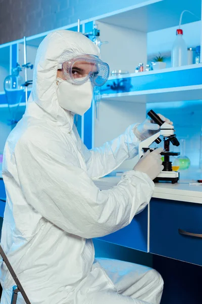 Científico en traje de materiales peligrosos y gafas sentado cerca del microscopio en el laboratorio - foto de stock