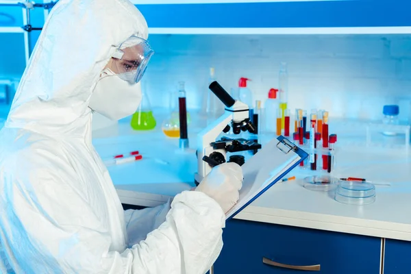 Científico en traje de materiales peligrosos y gafas escribiendo cerca del microscopio - foto de stock