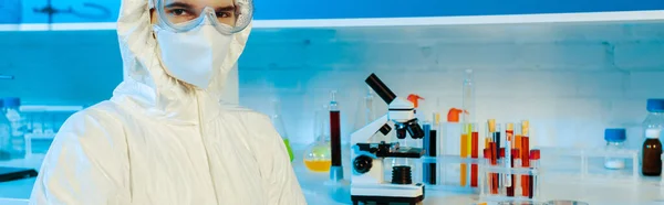 Панорамный снимок ученого в защитном костюме и очках возле микроскопа — стоковое фото