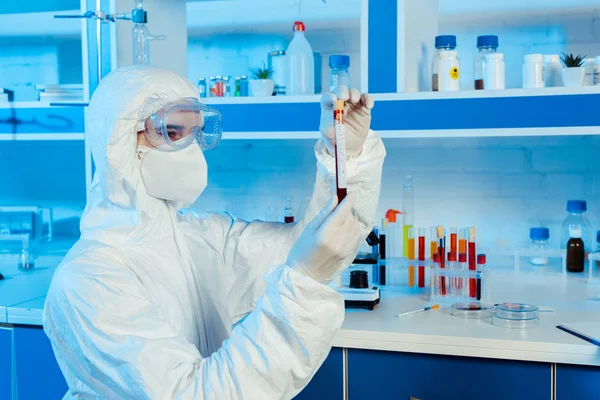 Científico en traje de materiales peligrosos y guantes de látex que sostiene el tubo de ensayo con letras de coronavirus - foto de stock