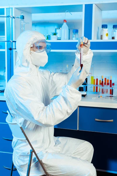 Científico en traje de materiales peligrosos y guantes de látex que sostiene el tubo de ensayo con letras de muestra y coronavirus - foto de stock