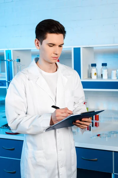 Handsome scientist in white coat holding clipboard and pen in laboratory — Stock Photo