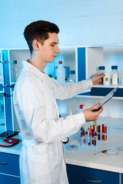 Side view of handsome scientist touching bottle with toxic sign and looking at clipboard — Stock Photo