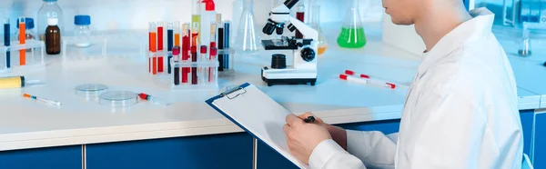 Panoramic shot of scientist in white coat holding clipboard and pen in laboratory — Stock Photo