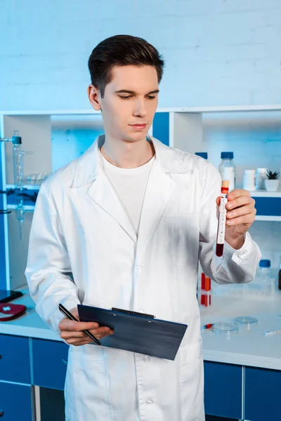 Handsome scientist holding test tube with coronavirus lettering and clipboard — Stock Photo