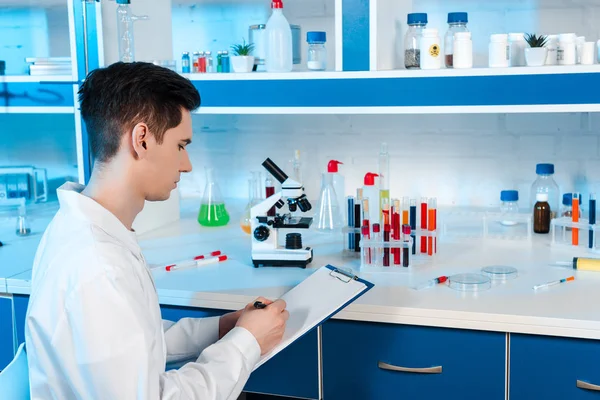 Guapo científico escribiendo cerca de microscopio y tubos de ensayo - foto de stock