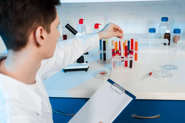 Foco seletivo do cientista no revestimento branco segurando prancheta e tocando tubo de ensaio — Fotografia de Stock