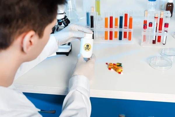 Selective focus of scientist in latex gloves holding bottle with danger sign near pills — Stock Photo