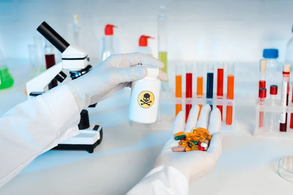 Обрезанный вид ученого в латексных перчатках, держащих бутылку с опасным знаком и таблетками — стоковое фото
