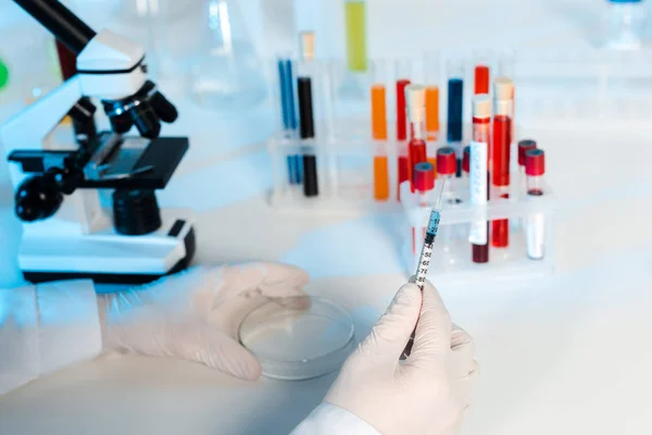 Cropped view of scientist in latex gloves holding glass test plate and syringe — Stock Photo